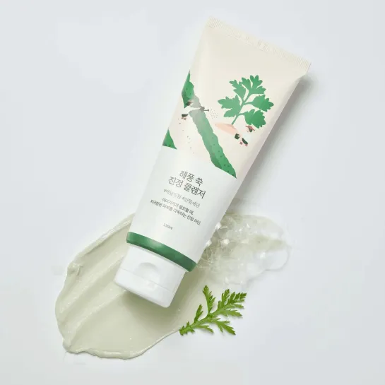 Korean skincare cleanser tube with green leaf design, creamy texture swatch, and fresh herb on white background
