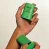 A close up of a hand applying A green haruharu wonder black bamboo daily soothing sun shield sun stick SPF 50+ PA++++ with very high protection and 20g weight on a white background.